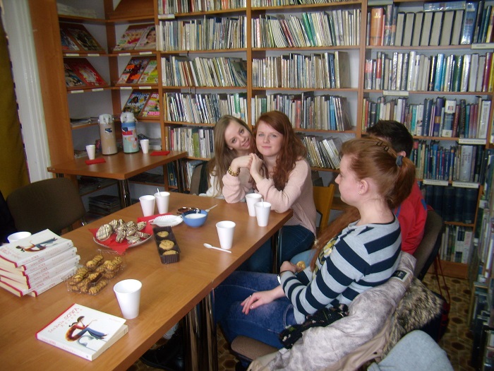 Wnętrze biblioteki, przy stoliku siedzą członkinie DKK wspólnie omawiają  książkę Izabeli Sowy "Agrafka".