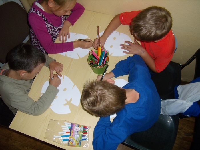Przy stoliku siedzi czworo dzieci wspólnie kolorują maski.