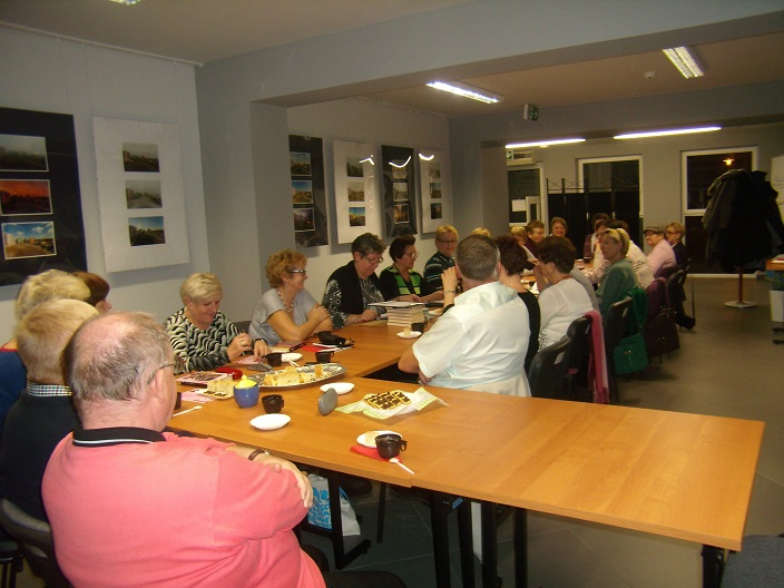 Wnętrze biblioteki,mediateka. Przy stolikach siedzą członkowie DKK UTW wspólnie omawiają książkę Beaty Pawlikowskiej "Blondynka Tao", na stolikach kawa i mały poczęstunek. 