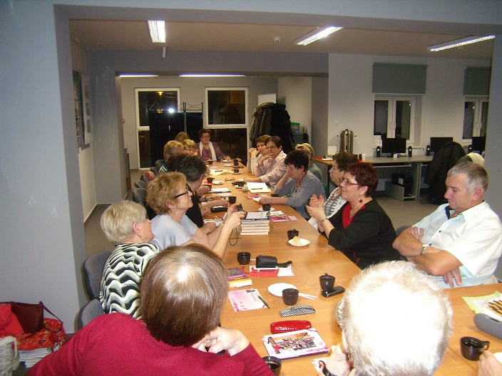 Wnętrze biblioteki,mediateka. Przy stolikach siedzą członkowie DKK UTW wspólnie omawiają książkę Beaty Pawlikowskiej "Blondynka Tao", na stolikach kawa i mały poczęstunek. 