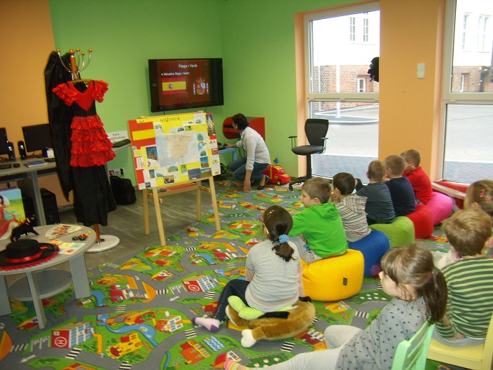 Na kolorowych pufach siedzą dzieci wspólnie biorą udział w lekcji bibliotecznej na temat kultury Hiszpanii.
