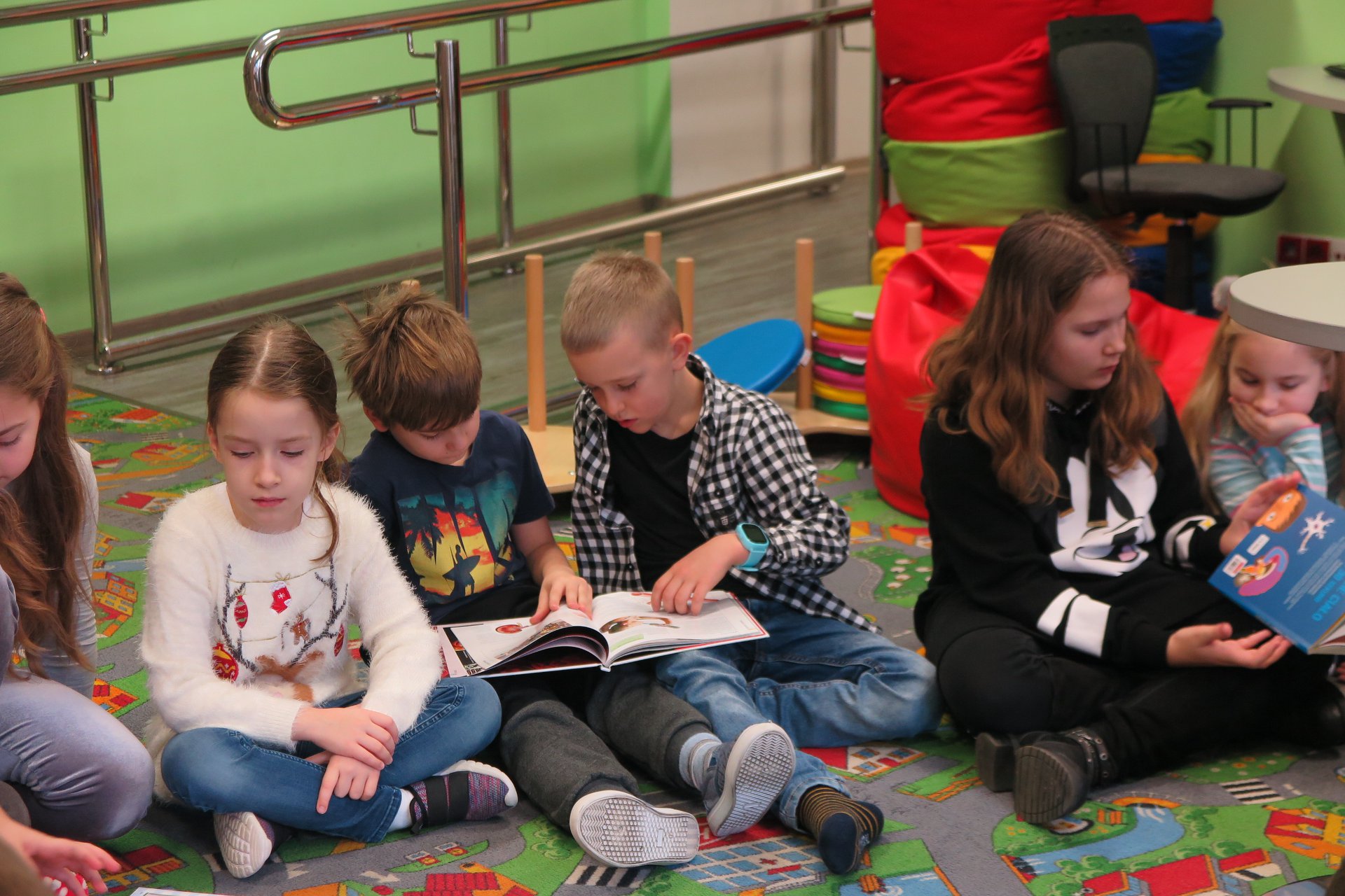 Wnętrze biblioteki (Klub Malucha) sześcioro dzieci siedzi na podłodze dwoje z nich czyta książkę.