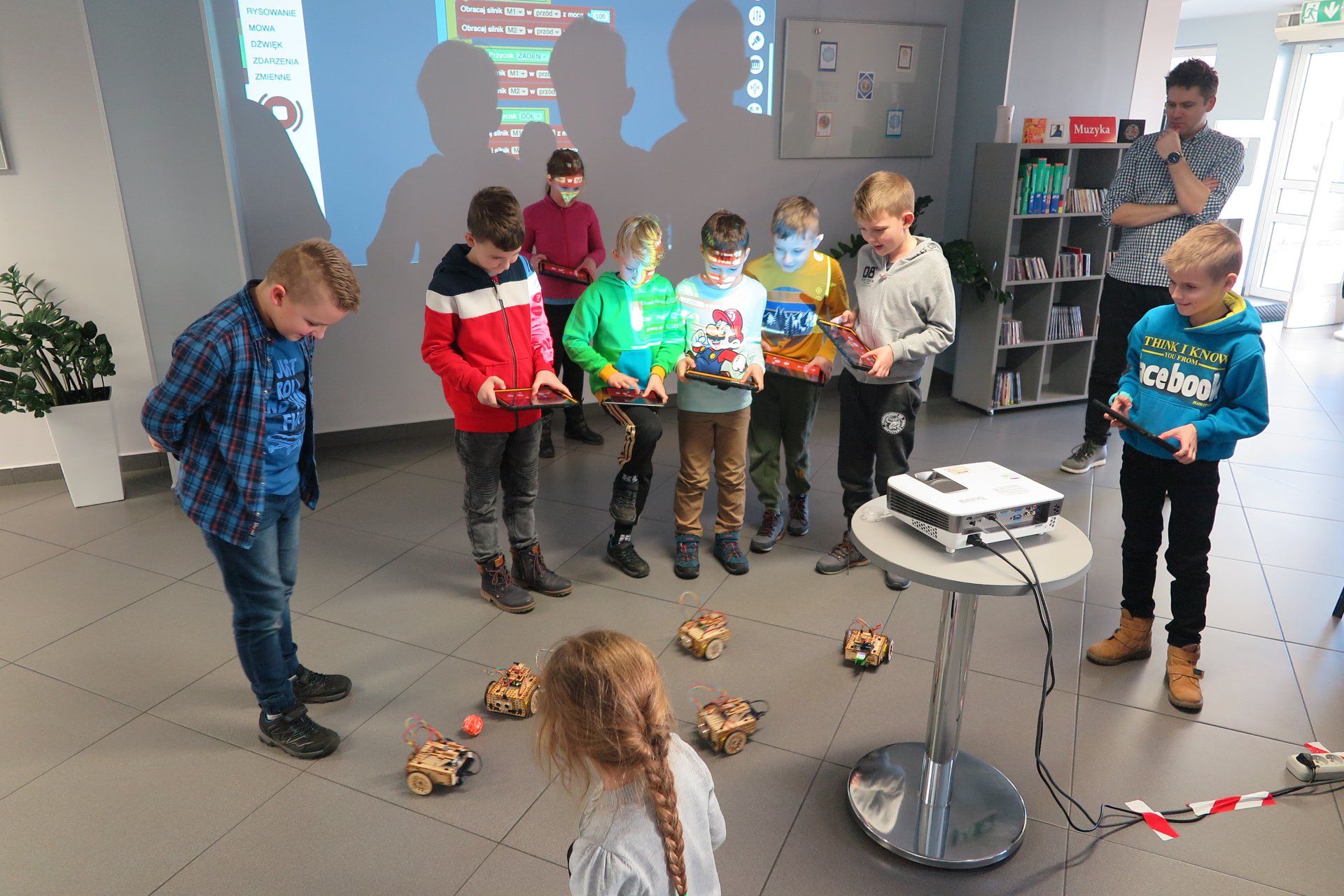 Wnętrze biblioteki (mediateka) zajęcia z robotami prowadzi pan Paweł obok dzieci biorą udział w szkoleniu na podłodze jeżdżące roboty LOFI
