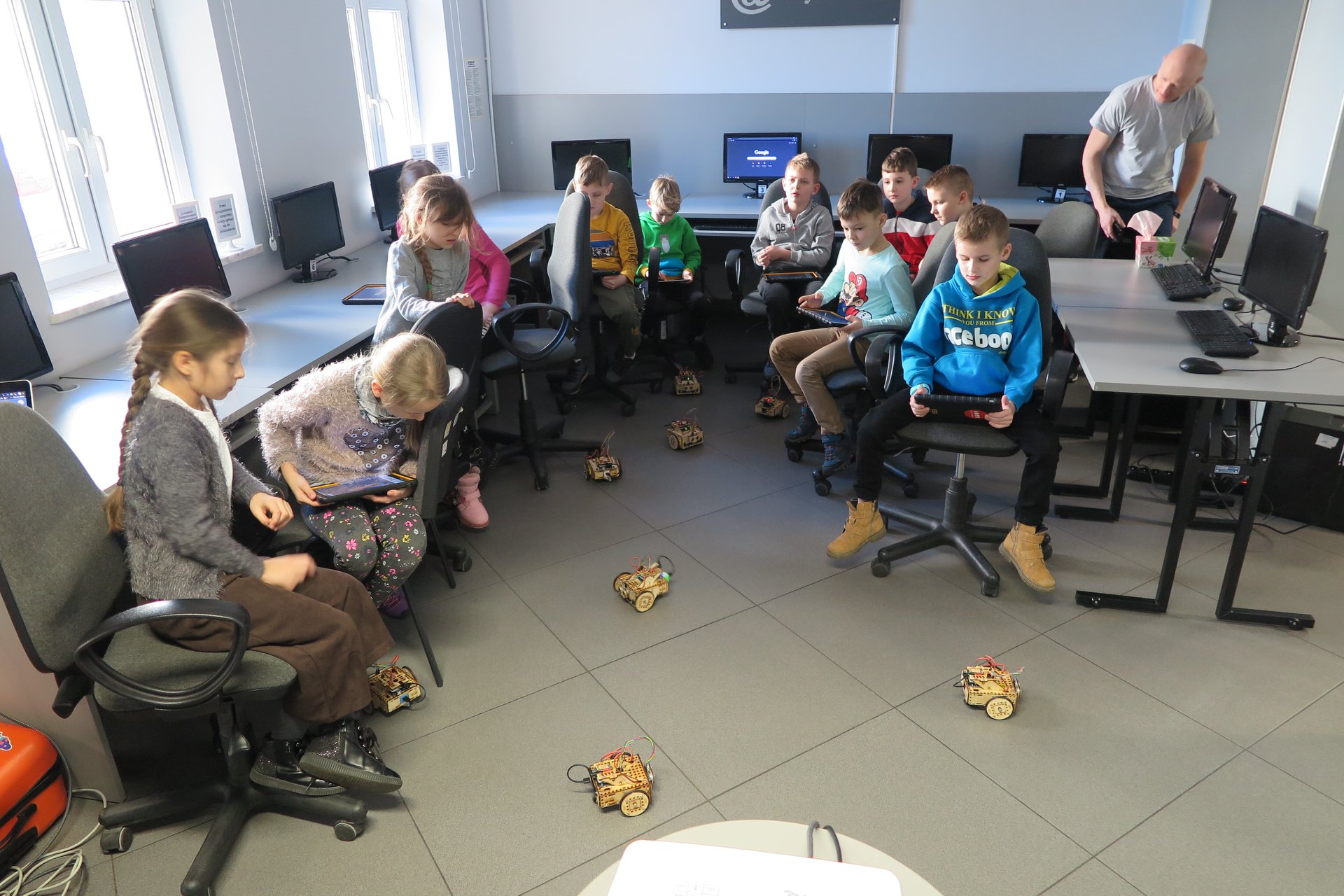 Wnętrze biblioteki (mediateka) zajęcia z robotami na krzesełkach siedzą dzieci, na podłodze poruszające się roboty. 