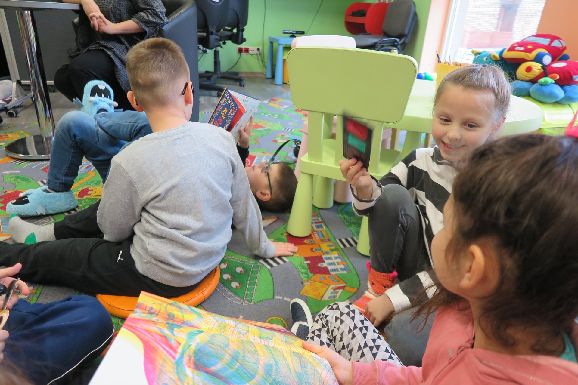 Wnętrze biblioteki (Klub Malucha) czworo dzieci bawi się na podłodze i czyta książki.