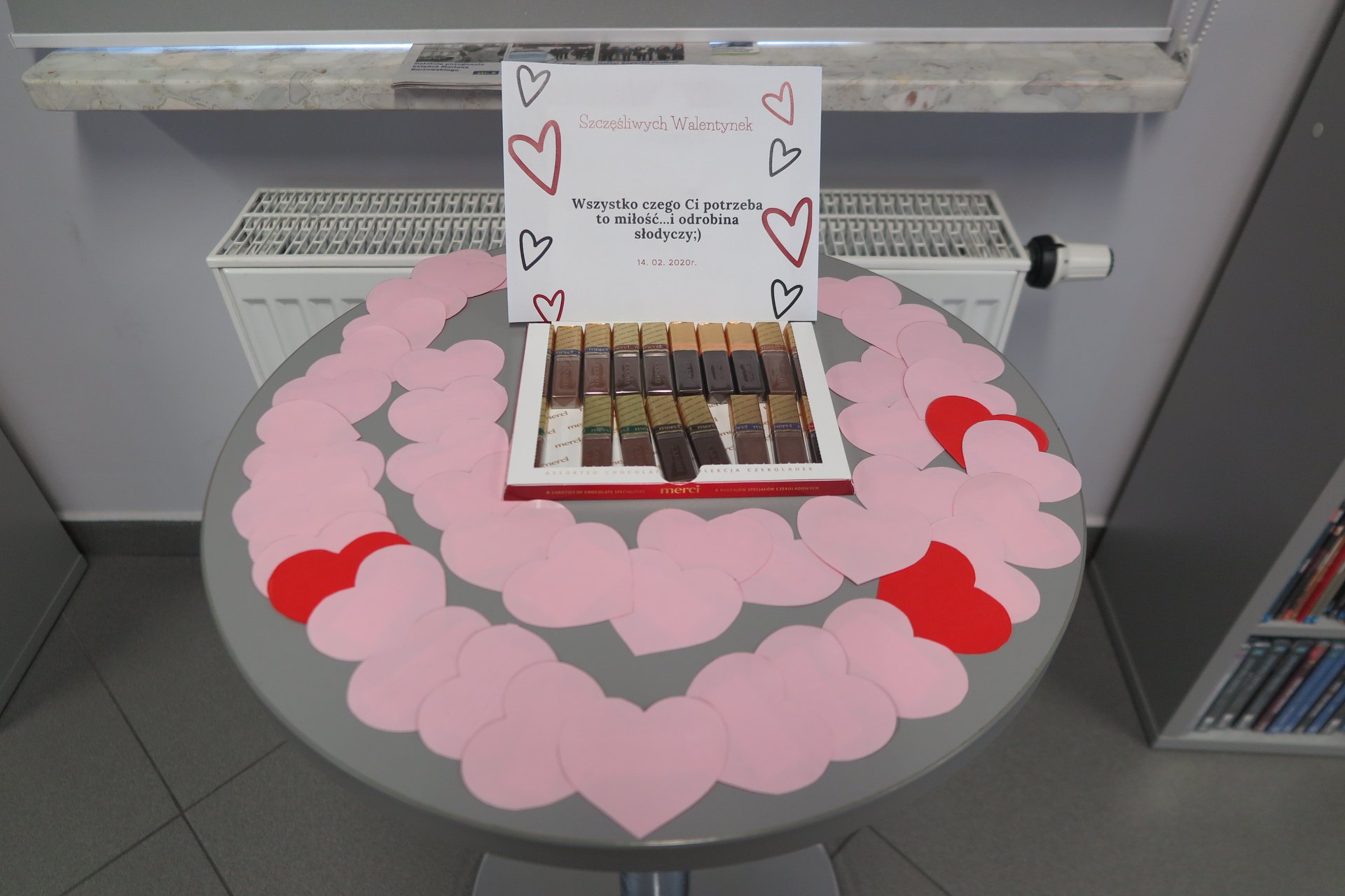 Okrągły stolik na nim wycięte z papieru serca, czekoladki merci i kartka z napisem ''Szczęśliwych Walentynek. Wszystko czego Ci potrzeba to miłość... i odrobina słodyczy :) ''.