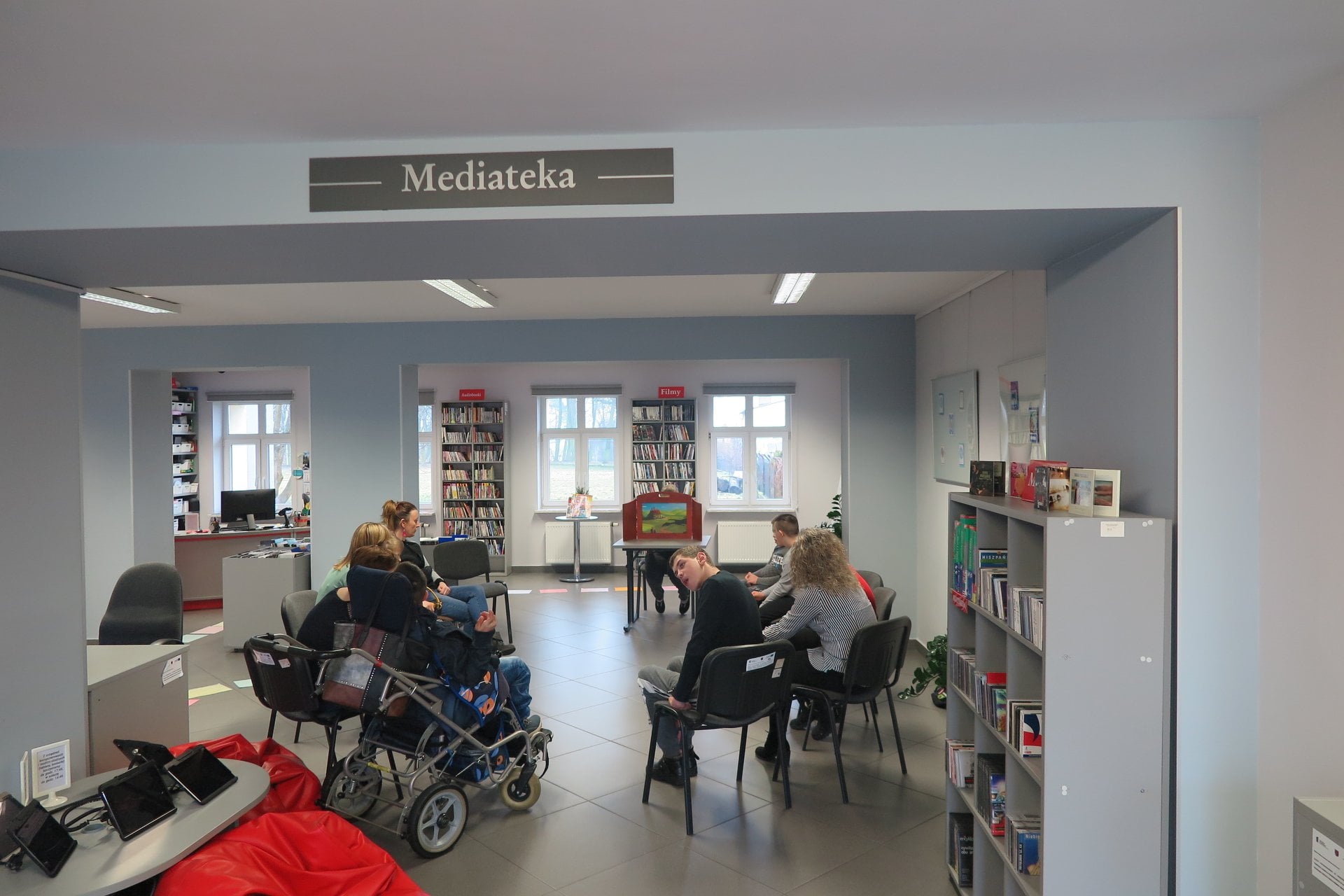 Wnętrze biblioteki, mediateka. Dzieci z Ośrodka Rehabilitacyjno-Edukacyjno- Wychowawczego z Działdowa wraz z opiekunami słuchają teatrzyku Kamishibai który czyta bibliotekarka Małgorzata Trąmpczyńska.