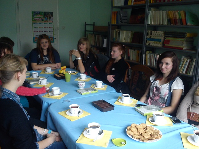 Przy stoliku siedzą członkowie DKK ZS Malinowo wspólnie omawiają książkę "Dziecko Ulicy", na stole kawa i ciastka.