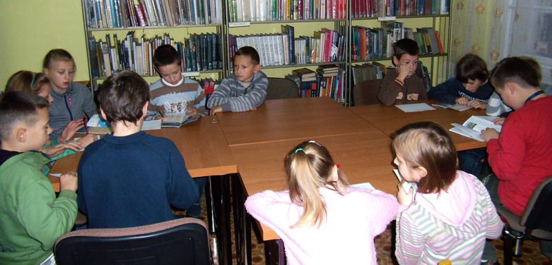 Wnętrze biblioteki, przy stolikach siedzą dzieci w tle regały z książkami. 