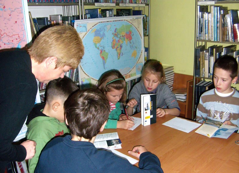 Wnętrze biblioteki, przy stolikach siedzą dzieci w tle regały z książkami oraz mapa świata.