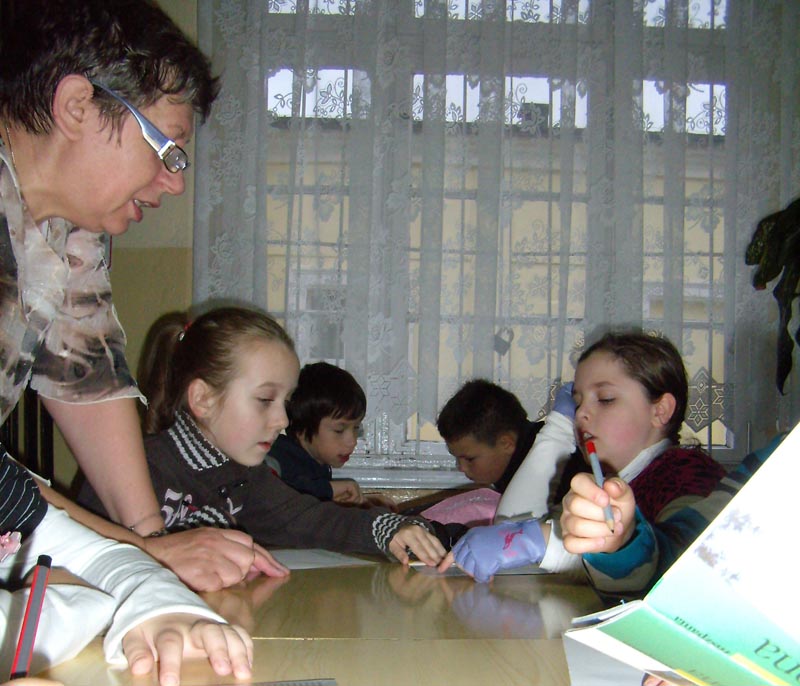 Wnętrze biblioteki, przy stolikach siedzą dzieci zajęcia prowadzi Ewa Sotomska.
