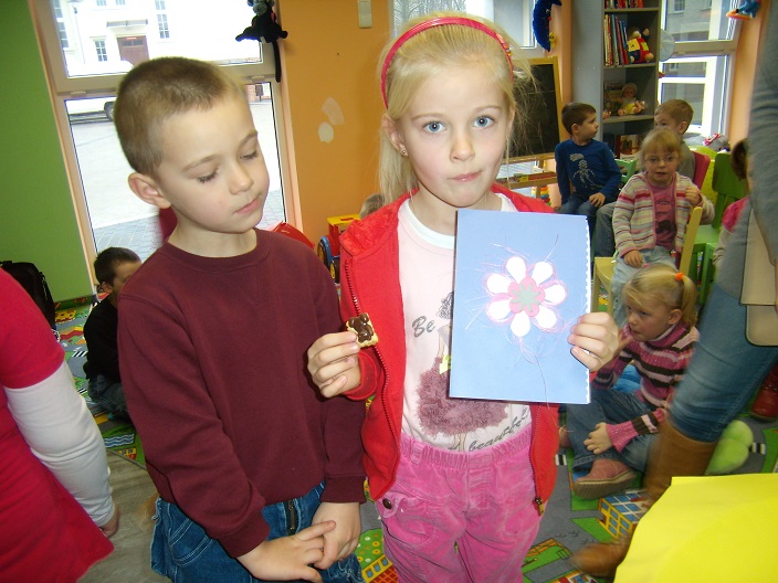 Dziewczynka trzyma w ręku prace plastyczną biały kwiatek obok niej stoi chłopiec.
