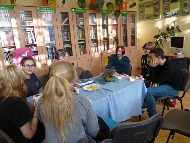 Wnętrze biblioteki, przy stoliku siedzi pani Ewa Sotomska i członkinie DKK wspólnie omawiają książkę Grzegorza Gortata „Miasteczko ostatnich westchnień”.