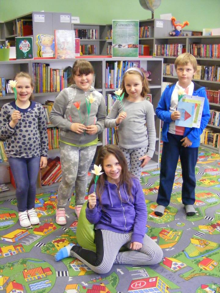 Przy regale z książkami stoi pięcioro dzieci z papierowymi tulipanami.