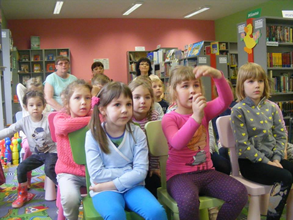Na krzesełkach siedzą dzieci, biorą udział w zajęciach z bibliotekarką Anią.