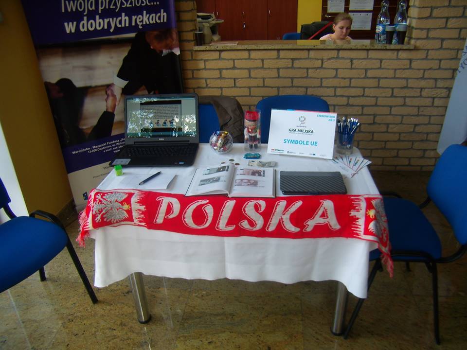 Na stoliku leży tablet, laptop, książka i plakat z napisem Gra Miejska Symbole UE.