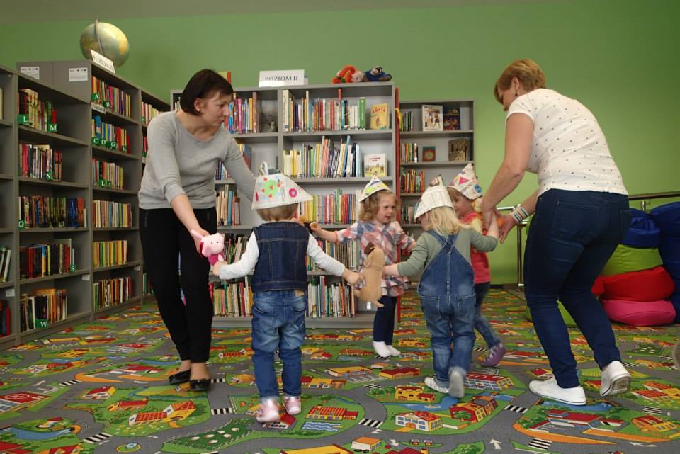 Czworo dzieci z opiekunką i bibliotekarką Anią podczas zabawy w tle regały z książkami.