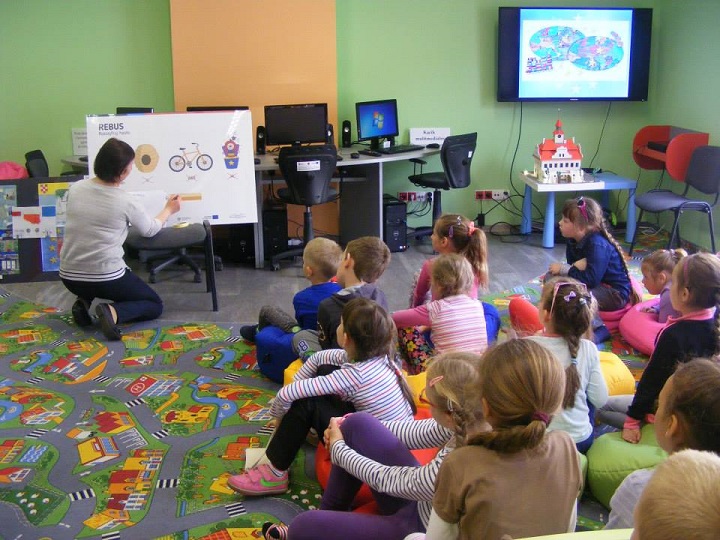 Grupa dzieci siedzi na kolorowych pufach, bibliotekarka Ania prowadzi lekcję na temat Unii Europejskiej w tle telewizor z prezentacją.
