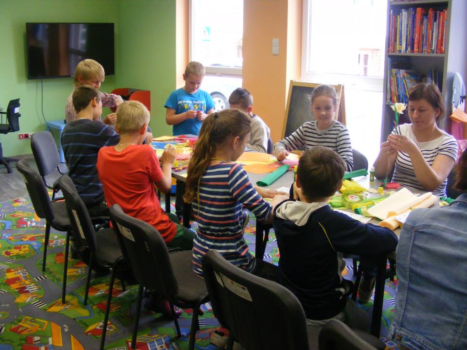 Wnętrze biblioteki, przy stoliku siedzą dzieci z bibliotekarką Anią razem wykonują prace plastyczne. 