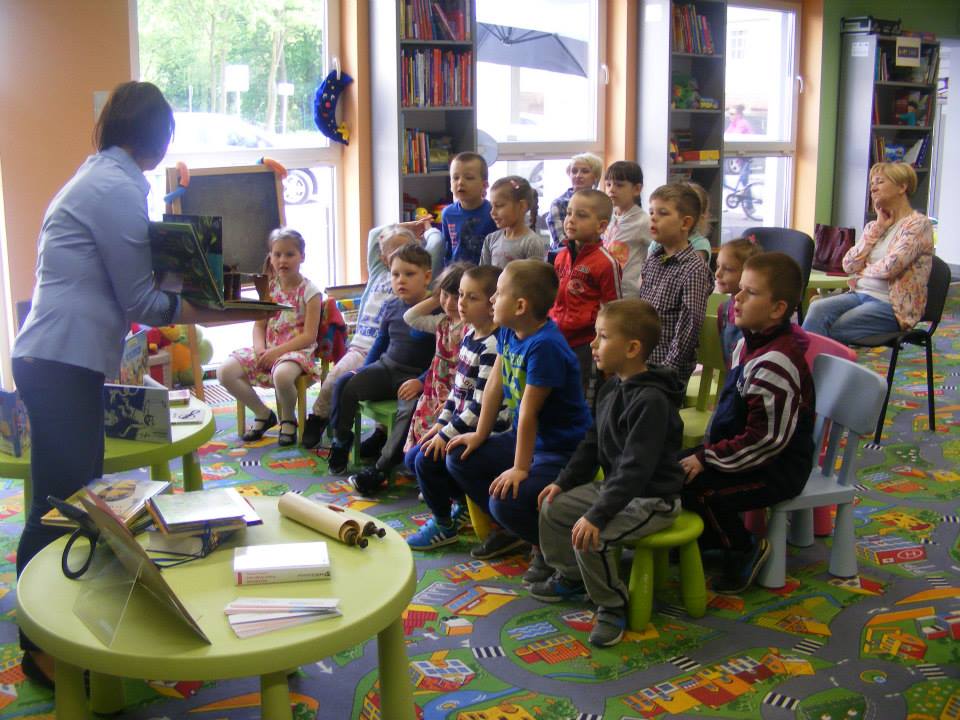 Wnętrze biblioteki, grupa dzieci siedzi na krzesełkach, bibliotekarka Ania prowadzi zajęcia na temat książki.