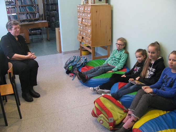 Wnętrze biblioteki, pani Ewa Sotomska i grupa dzieci z DKK wspólnie omawiają  książkę Beaty Ostrowickiej " Jest taka historia".