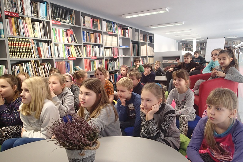 Wnętrze biblioteki, grupa dzieci siedzi na fotelach bierze udział w warsztatach z bajką a prowadzi je bajkopisarz Marek Samselski po lewej stronie regał z książkami.