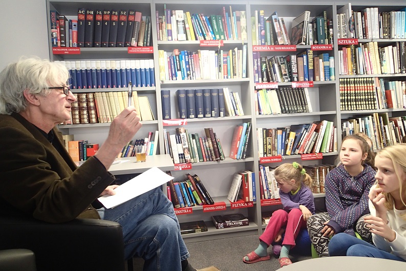Na fotelu siedzi Marek Samselski prowadzi zajęcia dla dzieci na temat bajek, w tle regał z książkami.