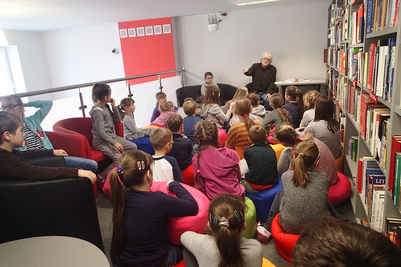 Wnętrze biblioteki, grupa dzieci siedzi na fotelach bierze udział w warsztatach z bajką a prowadzi je bajkopisarz Marek Samselski po prawej stronie regał z książkami.