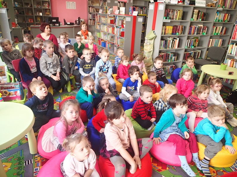 Grupa dzieci siedzi na kolorowych pufach, w tle regały z książkami.