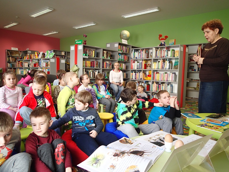Grupa dzieci siedzi na krzesełkach, lekcję na temat znaczenia jajka w życiu człowieka jako symbolu Wielkanocnego prowadzi bibliotekarka, w tle regały z książkami.