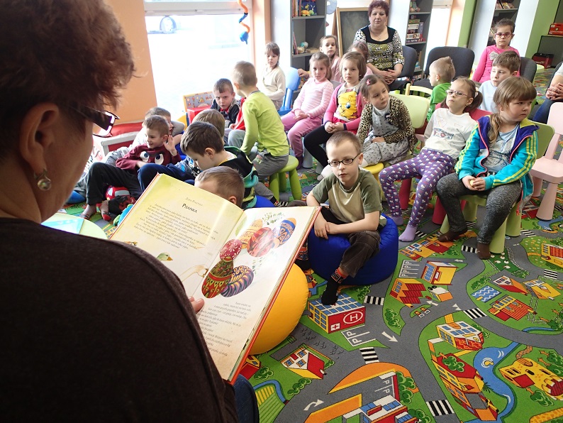 Grupa dzieci siedzi na krzesełkach, bibliotekarka czyta dzieciom książkę pt. ,,Pisanka''.