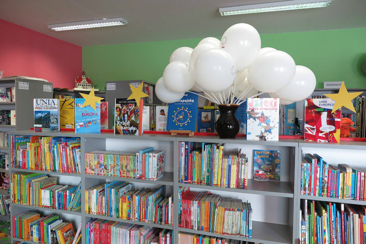 Wnętrze biblioteki, regały z książkami dla dzieci na górnej półce stoją w czarnym wazonie na piku białe baloniki.