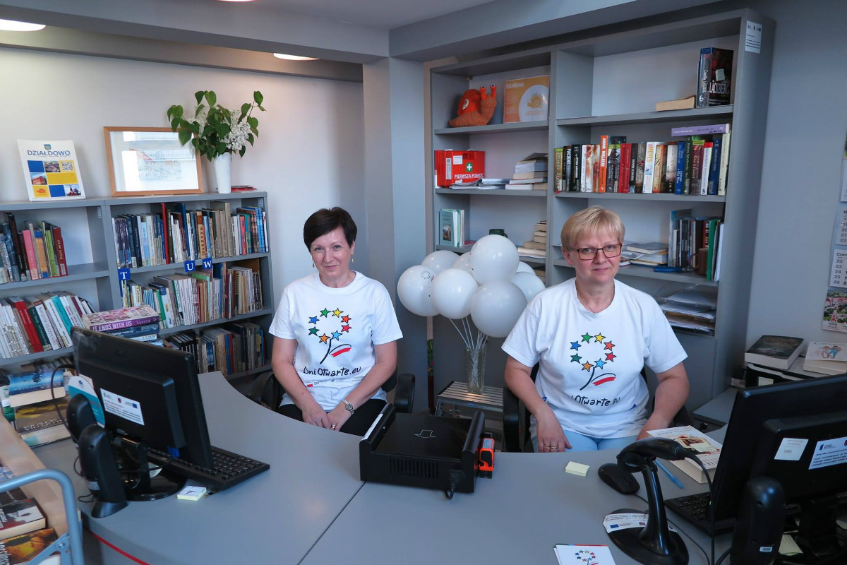 Wnętrze biblioteki, przy komputerach dwie bibliotekarki Pani Mariola Wilkowska i Elżbieta Wojcieszek w koszulkach z napisem ''Dni Otwarte Funduszy Europejskich''. 
