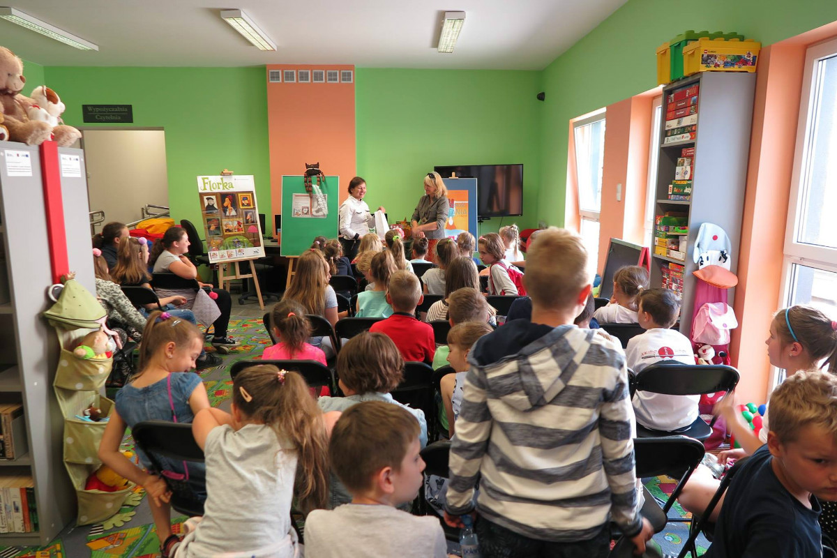 Wnętrze biblioteki, na krzesełkach siedzą dzieci biorą udział w spotkaniu z Roksaną Jędrzejewską-Wróbel autorką książek dla dzieci m.in. ''Florka''. W tle trzy sztalugi z plakatami.