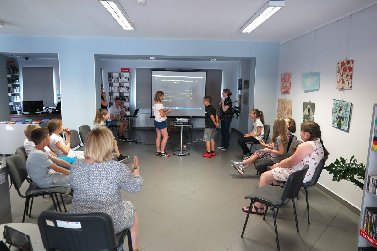 Wnętrze biblioteki, w kole siedzi młodzież biorąca udział w  w „Familiadzie Europejskiej”, w tle monitor z prezentacją z wiedzy o Unii Europejskiej.