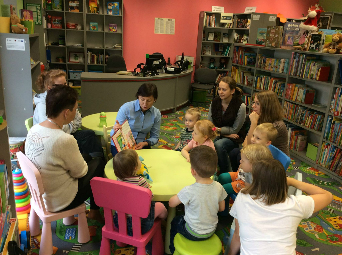 Wnętrze biblioteki. Przy stoliku siedzą dzieci z opiekunami, bibliotekarka Ania Wiśniewska czyta opowiadanie "Boże Narodzenie Tupcia Chrupcia". 