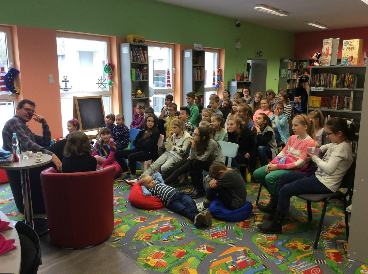 Wnętrze biblioteki, na krzesełkach siedzą dzieci biorą udział w spotkaniu w ramach DKK z Pawłem Beręsewiczem.