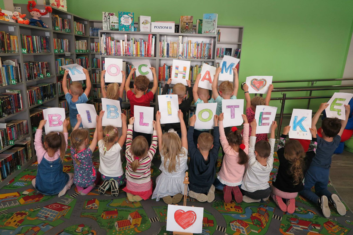Wnętrze biblioteki. Dzieci trzymają w górze kolorowe literki, które wcześniej narysowały, razem utworzyły napis ''Kocham Bibliotekę''. 