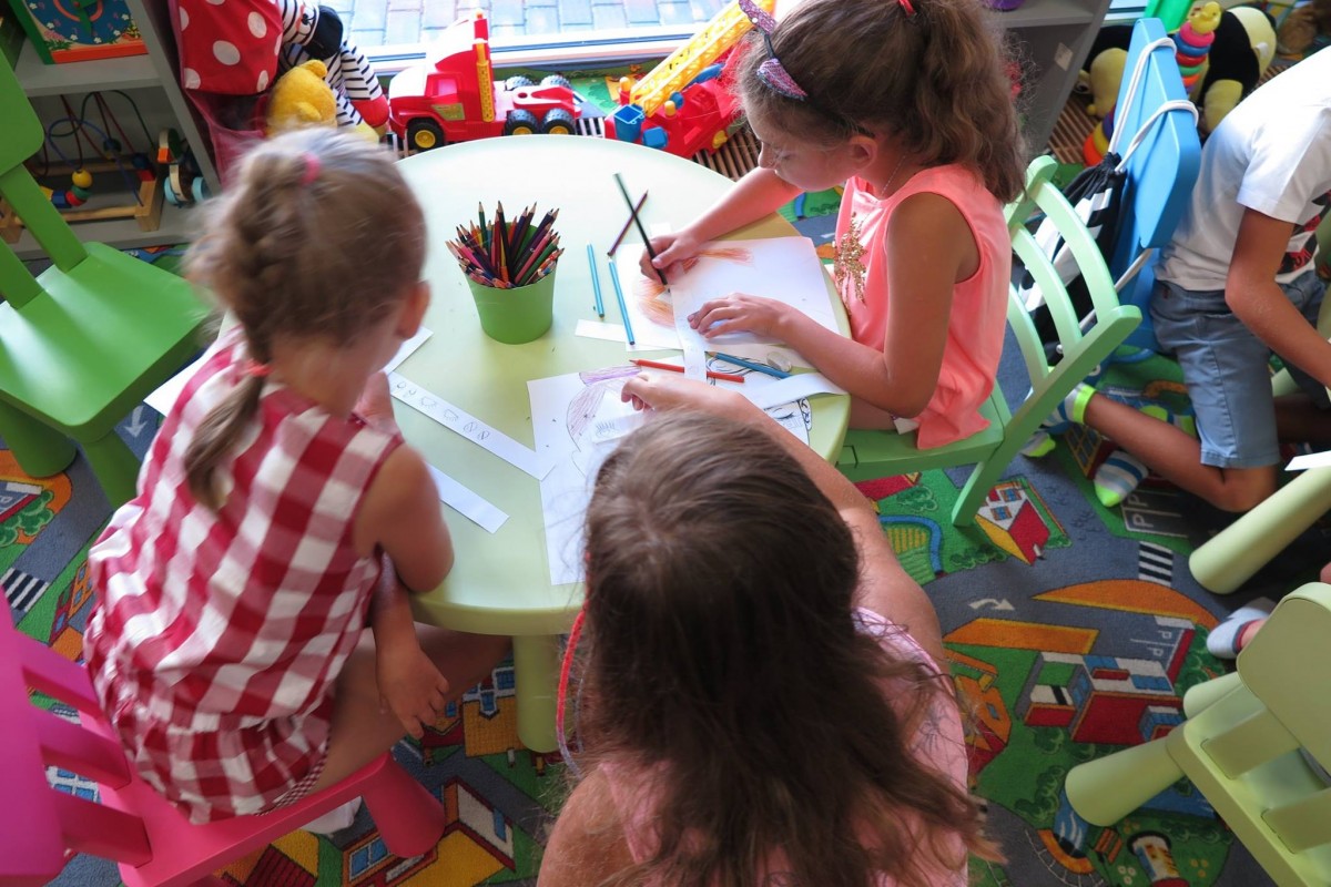Przy stoliku siedzą trzy dziewczynki i rysują na białych kartkach. 