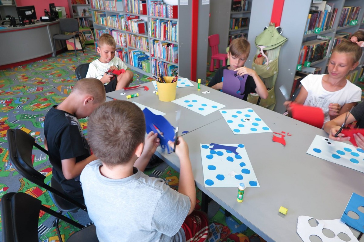 Wnętrze biblioteki. Przy stolikach siedzą dzieci i wycinają z kolorowego papieru kraby. 