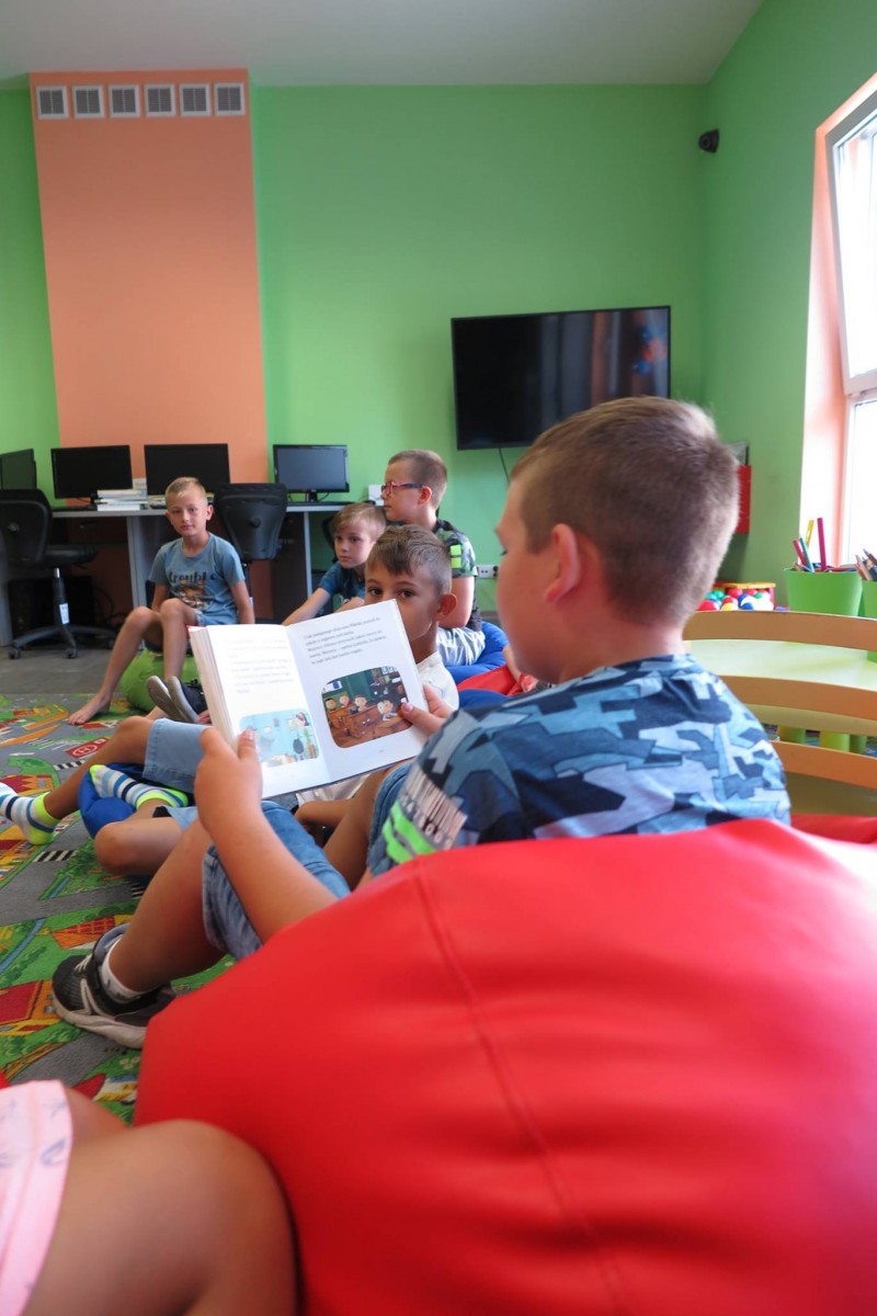 Wnętrze biblioteki. Na pufach siedzą dzieci, jeden z chłopców czyta  o kolejnych przygodach Mikołajka i jego kolegów. 