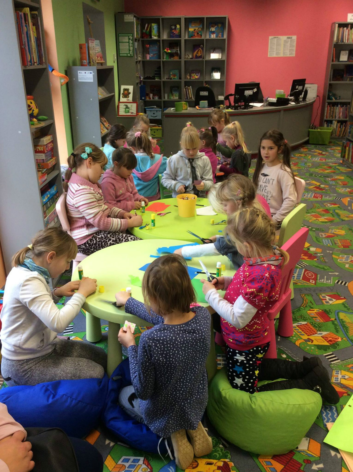 Wnętrze biblioteki, przy stolikach siedzą dzieci razem wykonują prace plastyczne.