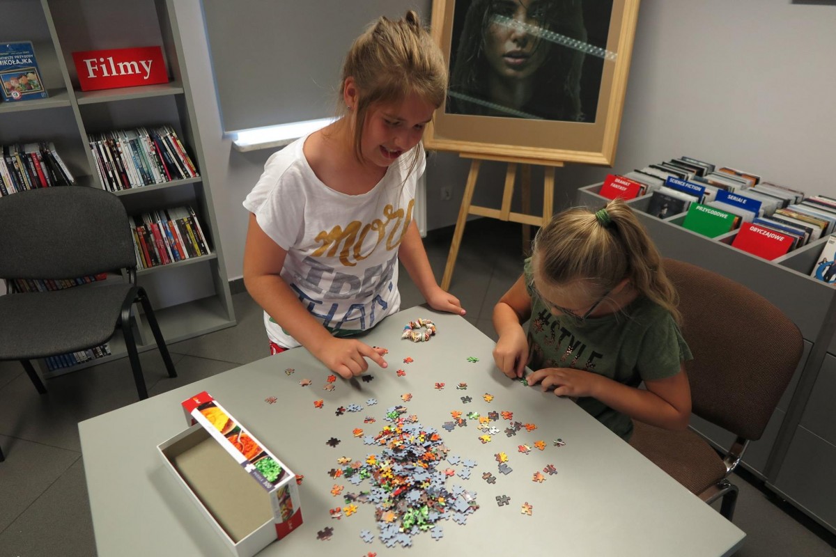 Wnętrze biblioteki,mediateka. Przy stoliku dwie dziewczynki układają puzzle na czas.