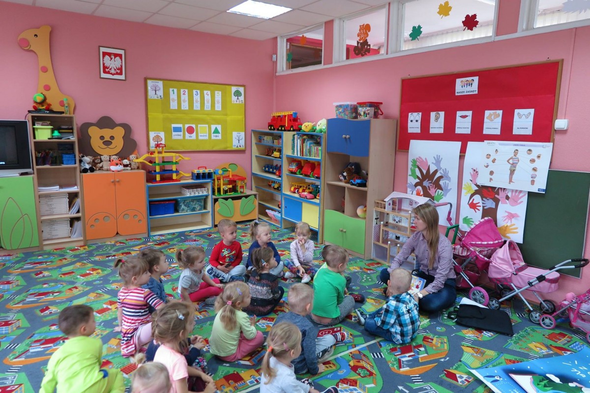 Wnętrze Przedszkola nr w Działdowie. Na kolorowej wykładzinie siedzą dzieci i słuchają teatrzyku paluszkowego "Trzy świnki", czyta bibliotekarka Ola Cybulska.