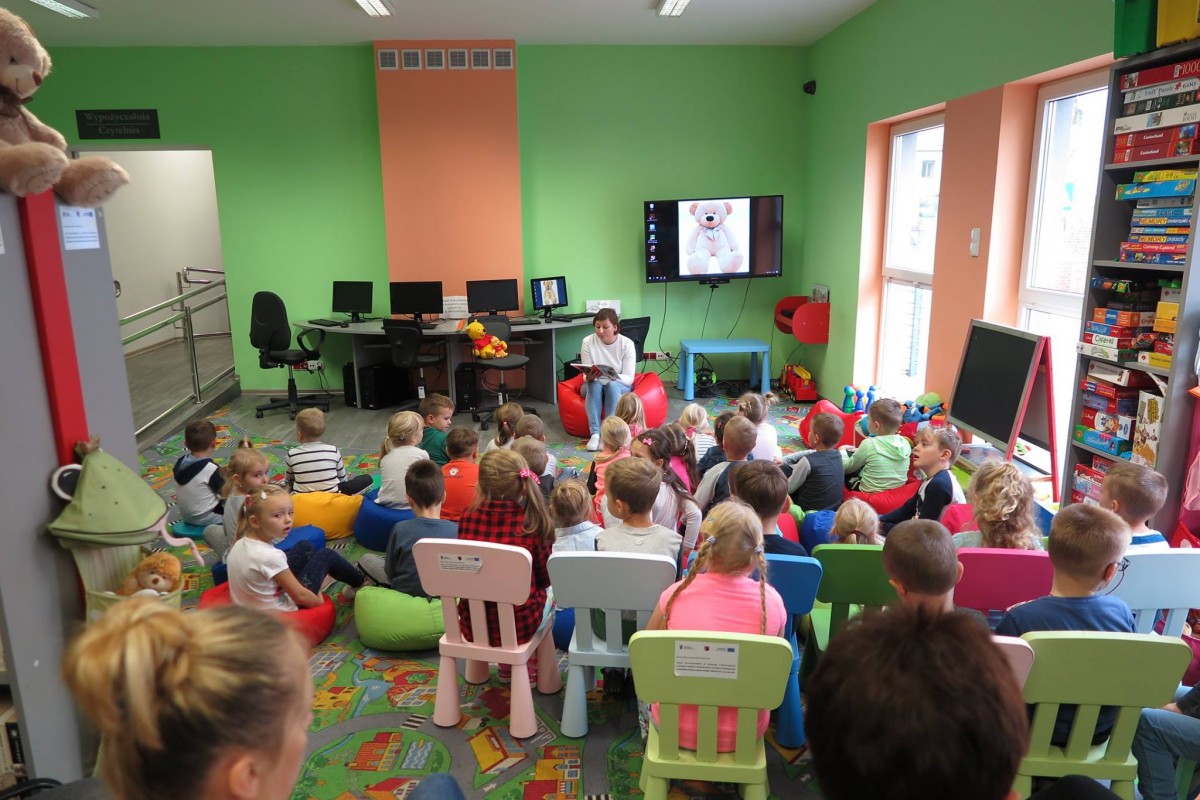 Wnętrze biblioteki. Na krzesełkach i kolorowych pufach siedzą dzieci, słuchają opowiadania o Misiu Uszatku.