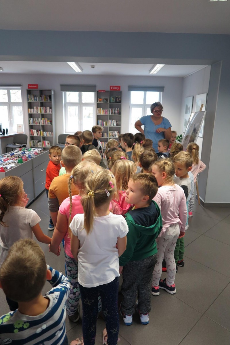 Wnętrze biblioteki. Dzieci zwiedzają oddział mediateki. Opowiada o nim bibliotekarka Dorota Kusik. 