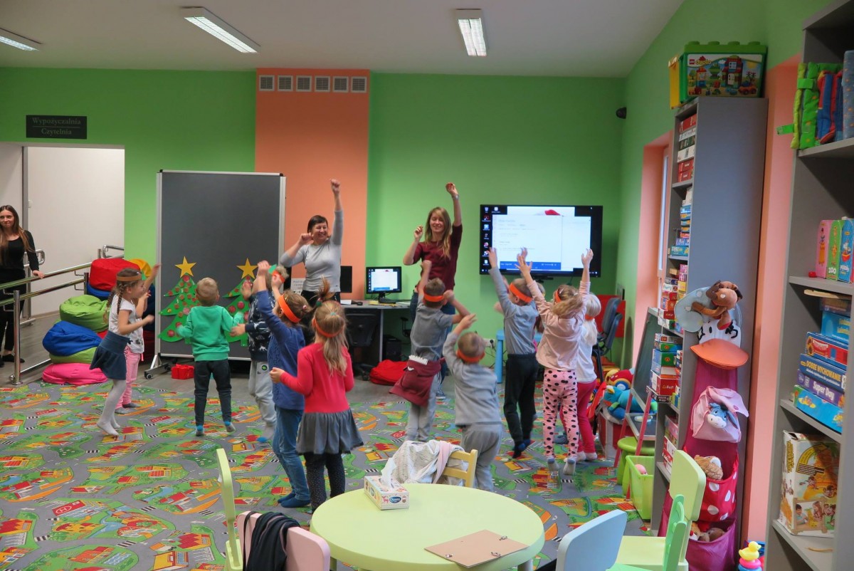 Wnętrze biblioteki. Grupa dzieci z Przedszkola "U Misia" uczestniczą w zabawie prowadzonej przez dwie bibliotekarki Anię Wiśniewską i Aleksandrę Cybulską.