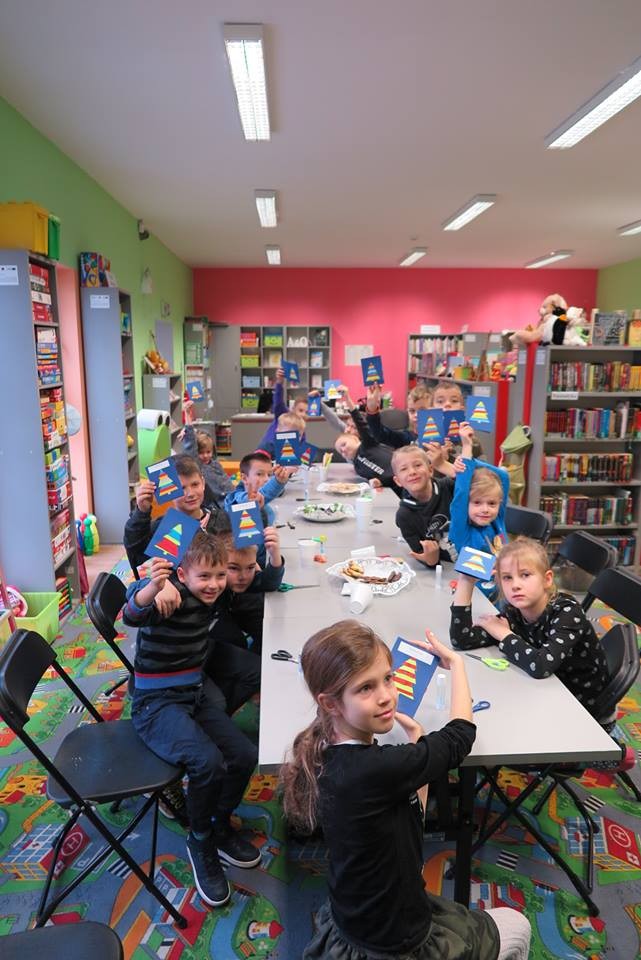 Wnętrze biblioteki. Przy stolikach siedzą dzieci z DKK SP nr 1 i trzymają w górze zrobione przez siebie kartki świąteczne dla swoich bliskich. 
