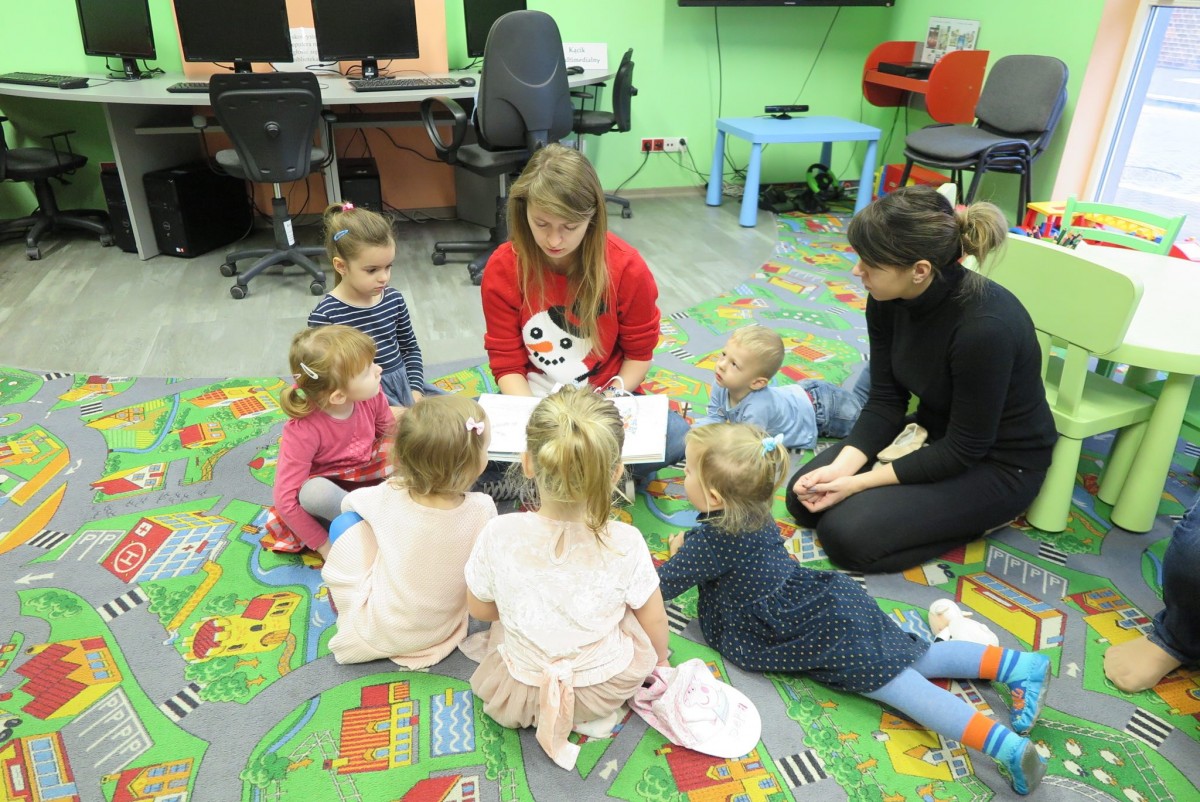 Wnętrze biblioteki. Na kolorowej wykładzinie siedzą dzieci z Klubu Malucha i słuchają jak czyta bibliotekarka Aleksandra Cybulska książkę "Prezent dla Świętego Mikołaja".