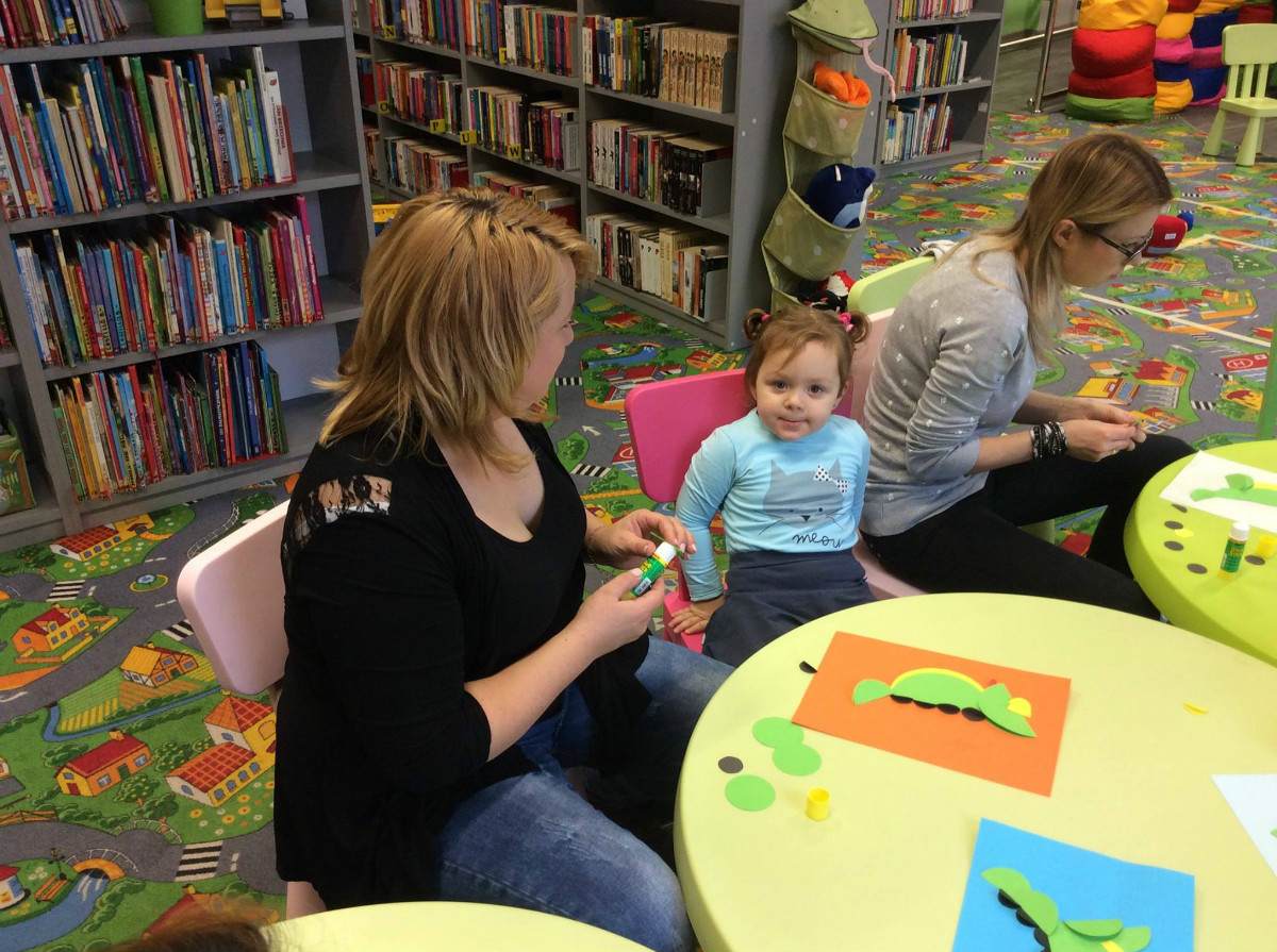 Wnętrze biblioteki, przy stoliku siedzą dzieci z opiekunkami razem wykonują z papieru zielone krokodyle.